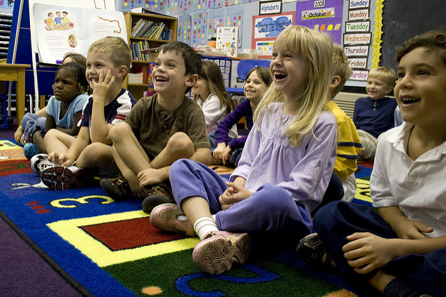12 Kinder von ungefähr fünf Jahren die auf einem bunten Teppich in einem Kindergarten sitzen und sich köstlich amüsieren