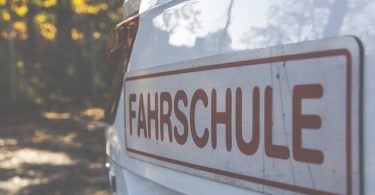 Nahaufnahme der hinteren Teils eines weissen Fahrschulautos auf dem ein großer Aufkleber mit roter Schrift klebt auf dem Fahrschule in Großbuchstaben steht