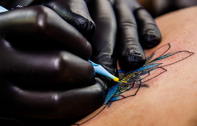 die Hände eines Tätowieres in schwarzen Gummihandschuhen wie er gerade eine blau-gelbe Lotusblüte tätowiert 