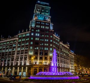 der Brunnen bei Nacht lila beleuchtet, links daneben ein gelb-schwarzes Taxi und dahinter das Generali Gebäude