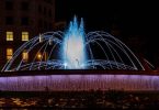 Der Brunnen bei Nacht bit weisser Beleuchtung man sieht die Lichter in den Fenstern des Gebäudes dahinter