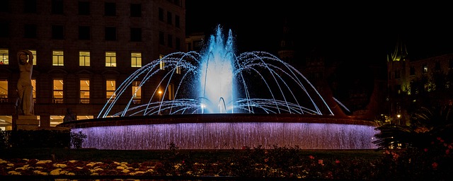 Der Brunnen bei Nacht bit weisser Beleuchtung man sieht die Lichter in den Fenstern des Gebäudes dahinter