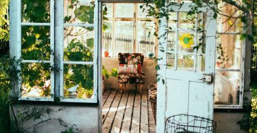Einblick in deine Wintergarten einer Wohnung, eine weisse Holztür , ein Sessel, viele Hängepflanzen und Holzboden umgeben von Fenstern durch die viel Licht reinscheint