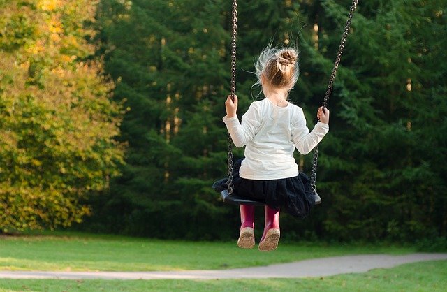 ein kleines blondes Mädchen mit roten Gummistiefeln auf einer Schaukel, das auf einen gelb-grünen Wald blickt