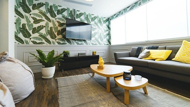 ein wohnzimmer mit einer grauen Couch mit gelben Kissen, dacor stehen ein kleiner und ein etwas größerer Holztisch, an der wand ein kleiner schwarzer Schrank und die Tapete ist weiß mit grünen Blättern