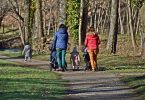 eine gepflasterter Weg der durch eine Park führt und auf dem zwei Frauen, eine mit blauer Jacke und die andere mit roter Jacke , jeweils einen Kinderwagen anschieben und zwischen ihnen und etwas weiter vor ihnen zwei kleine Kinder laufen