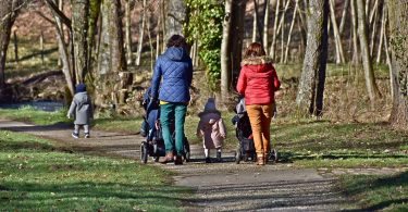 eine gepflasterter Weg der durch eine Park führt und auf dem zwei Frauen, eine mit blauer Jacke und die andere mit roter Jacke , jeweils einen Kinderwagen anschieben und zwischen ihnen und etwas weiter vor ihnen zwei kleine Kinder laufen