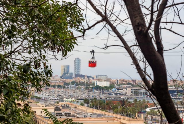 zwischen Baumästen ist über der Stadt Barcelona ein roter Wagon der Seilbahn zu sehen