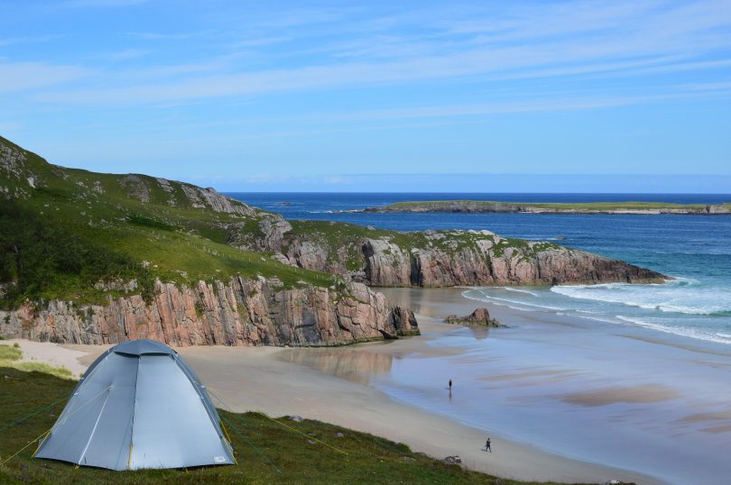 vorne links ein graues Zelt rechts Strand und Meer, grosse Klippen und zwei personen die am Strand entlang spazieren