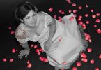 Eine junge Frau die auf einem schwarzen Fussboden mit roten Rosenblüten sitzt, sie hat ein weisses Hochzeitskleid an, die Haare hochgesteckt und bunte Sterne auf ihrer rechten Schulter tätowiert