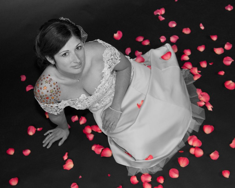 Eine junge Frau die auf einem schwarzen Fussboden mit roten Rosenblüten sitzt, sie hat ein weisses Hochzeitskleid an, die Haare hochgesteckt und bunte Sterne auf ihrer rechten Schulter tätowiert