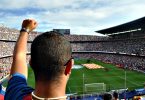 ein mann von hinten zu sehen der den linken arm hoch hält und seine Mannschaft von den zuschauertribühnen im Stadion anfeuert
