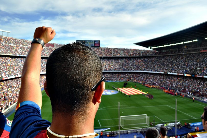 ein mann von hinten zu sehen der den linken arm hoch hält und seine Mannschaft von den zuschauertribühnen im Stadion anfeuert