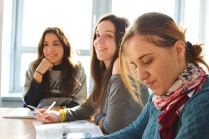 drei junge Frauen die an einem Tisch sitzen zwei lächeln nach vorne und eine schaut in ein Buch