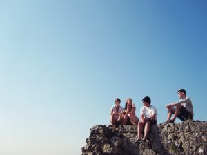 vier Jugendliche die auf einem Felsvorsprung sitzen danieder klarer blauer Himmel