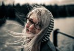 Eine junge Frau auf einer Brücke mit einer schwarz-weiss gestreiften Kapuze, langen blonden Harren und einer schwarzen Brille die in die Kamera lächelt