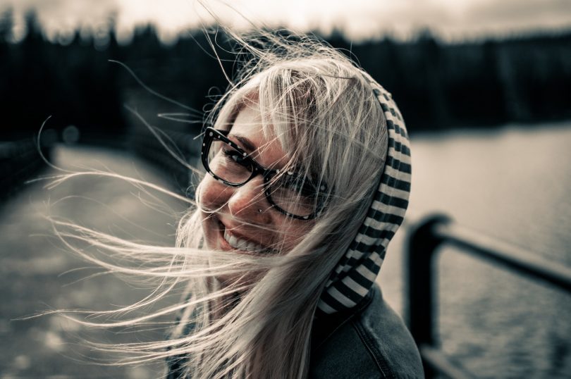 Eine junge Frau auf einer Brücke mit einer schwarz-weiss gestreiften Kapuze, langen blonden Harren und einer schwarzen Brille die in die Kamera lächelt