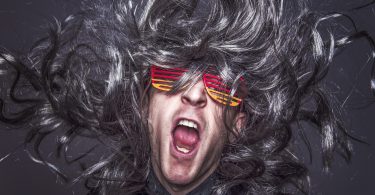 ein Mann mit dunkelgrauen Haaren, einer rot gelben Faschingsbrille und einem grauem Hemd der mit offenem Mund schreit