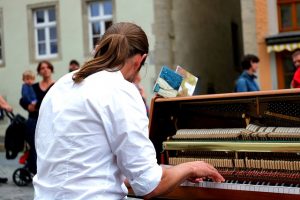 ein Mann der etwas auf einem Klavier mitten auf der Straße spielt, er hat lange braune Haare zu einem Pferdeschwanz zusammen gebunden und ein weisses Hemd an, man sieht ihn von Hinten und Leute um ihn herum