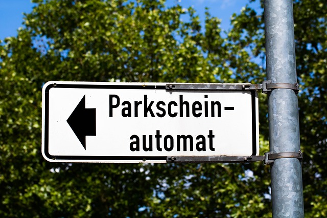 weisses Schild mit schwarzem Pfeil und geschrieben Parkscheinautomat, im Hintergrund Blätter eines Baumes