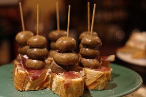 sechs Pintxos auf hellgrünem Teller: Baguette, Serrano Schinken und drei grüne Oliven