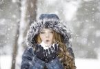 Frau mit langen lockigen Haaren, blauem Anorak und Handschuhen bläst aus ihrer Hand Schnee entgegen