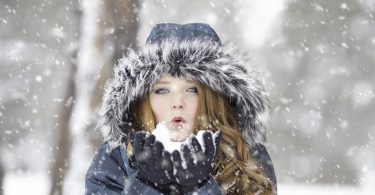 Frau mit langen lockigen Haaren, blauem Anorak und Handschuhen bläst aus ihrer Hand Schnee entgegen