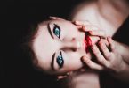 Frau mit hellblauen Augen, roten Lippen die leicht ihr Gesicht mit de Händen nach oben führt für das Foto