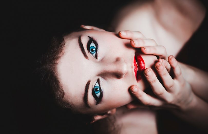 Frau mit hellblauen Augen, roten Lippen die leicht ihr Gesicht mit de Händen nach oben führt für das Foto