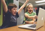 Ein Junge und ein Mädchen die vor Freude Jubeln und das Mädchen zeigt auf etwas auf dem Bildschirm eines grauen Apple Laptops