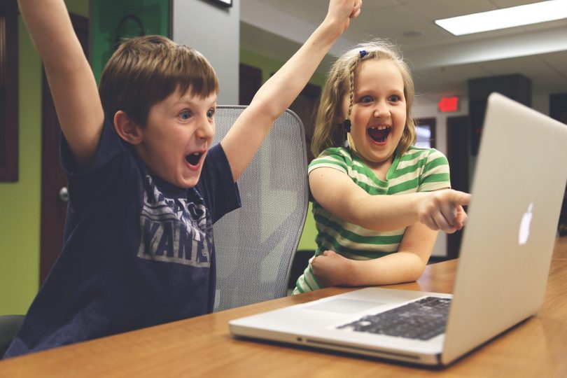 Ein Junge und ein Mädchen die vor Freude Jubeln und das Mädchen zeigt auf etwas auf dem Bildschirm eines grauen Apple Laptops