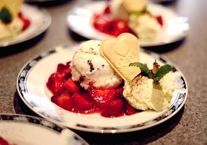 kleiner weisser Teller mit blauem Rand, darauf geschnittene Erdbeeren, zwei Kugeln Eis, Mintblätter und eine herzförmige Waffel