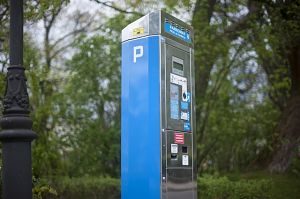 Ein blauer Parkautomat neben einer Laterne und grüne Bäume im Hintergrund