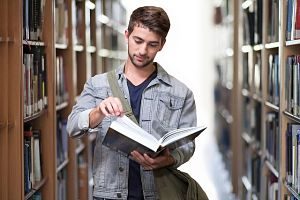 Ein junger Mann der gerade eine Seite eines grossen Buches umschlagen will während er durch eine Bibliothek läuft