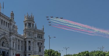 sieben Militärflugzeuge die in Dreiecks Formation die spanische Flagge mit Rauch über einem Rathaus überfliegen