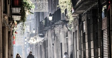 Fussgängerzone in Barcelona, ein Pärchen läuft unter der warmen Wintersonne die Gasse entlang, ein Teil der Weihnachtsbeleutung ist schon aufgehängt