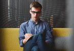 ein junger Mann mit schwarzer, eckiger Brille, Jeans und blauem Hemd sitzt auf einer gelben Bank und notiert etwas ein einem Heft