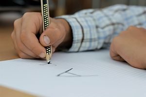 der arm eines Jungen mit einem blau-grau kariertem Hemd, er hält einen schwarzen Stift in der Hand und hat auf ein kariertes Blatt ein grosses und ein kleines a geschrieben