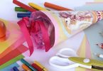 Eine kleine, rosten Schultüte auf einem weissen Tisch , drum herum liegen Scheren, Buntstifte, bunte Kartons etc