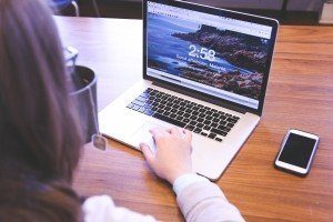 Frau die an einem Holztisch sitzt eine Tasse Tee in der linken Hand hält und mit der rechten Hand an einem Laptop arbeitet