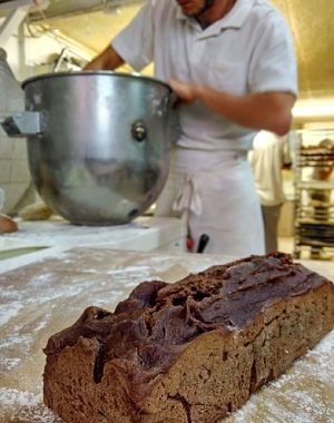 im Vordergrund ein dunkles rechteckiges Brot, im Hintergrund ein Bäcker der gerade teig in einer grossen Schüssel knetet