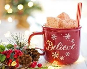 eine rote Tasse auf der in weiss believe steht mit mehreren Schneeflocken drumherum, ein weiss-rotes Röhrchen und weihnachtliche Dekoration neben der Tasse wie ein kleiner Tannenzweig, Misteln, Tannenzapfen und Lichter 