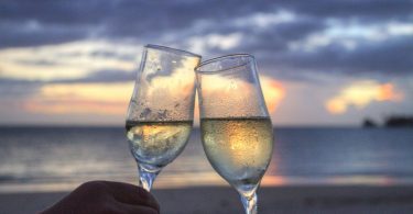 zwei Sektgläser die miteinander anstossen im Hintergrund das Meer , Wolken und ein Sonnenuntergang