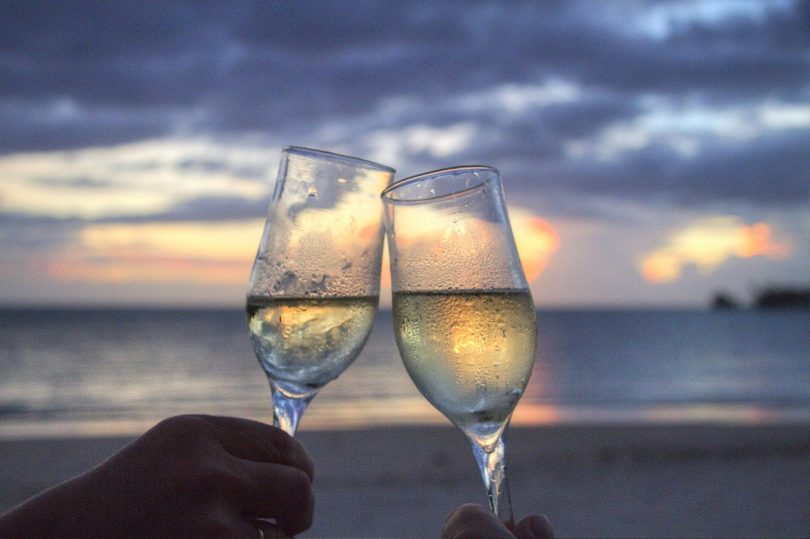 zwei Sektgläser die miteinander anstossen im Hintergrund das Meer , Wolken und ein Sonnenuntergang