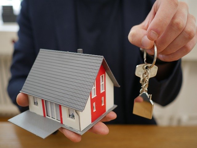 ein Mann in blauem Hemd der in seiner rechten Hand ein kleines rot-graues Haus hält und mit der linken Hand einen Schlüssel überreicht