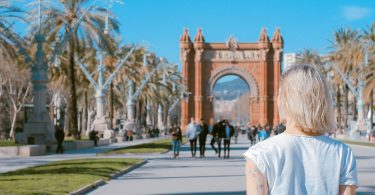 Die Allee zum Triumph Tor in Barcelona, es ist eine junge blonde Frau mit weisem T-shirt von hinten zu sehen, die auf einem Arm eine geometrische Figur und auf dem anderen Blumenzweige tätowiert hat