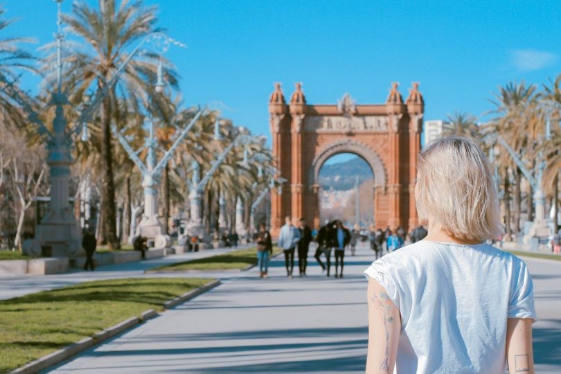 Die Allee zum Triumph Tor in Barcelona, es ist eine junge blonde Frau mit weisem T-shirt von hinten zu sehen, die auf einem Arm eine geometrische Figur und auf dem anderen Blumenzweige tätowiert hat
