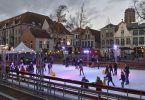 eine Schlittschuhbahn auf einem Marktplatz mit vielen Leuten darauf, Kinder mit Fahrhilfen und weihnachtslichter über der Bahn und in den bäumen