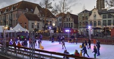 eine Schlittschuhbahn auf einem Marktplatz mit vielen Leuten darauf, Kinder mit Fahrhilfen und weihnachtslichter über der Bahn und in den bäumen