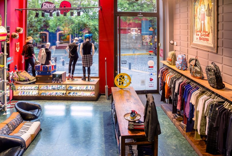 Innenraum eines tranigem modernen laden, rechts sind t-shirts, Jacken und Hemden aufgehängt am Eingang steht ein länglicher Holztisch, im Schaufenster vier Mannequin auf einem Regal in dem Schuhe eingeordnet sind und über den t-scirts verschiedenen Rucksäcke